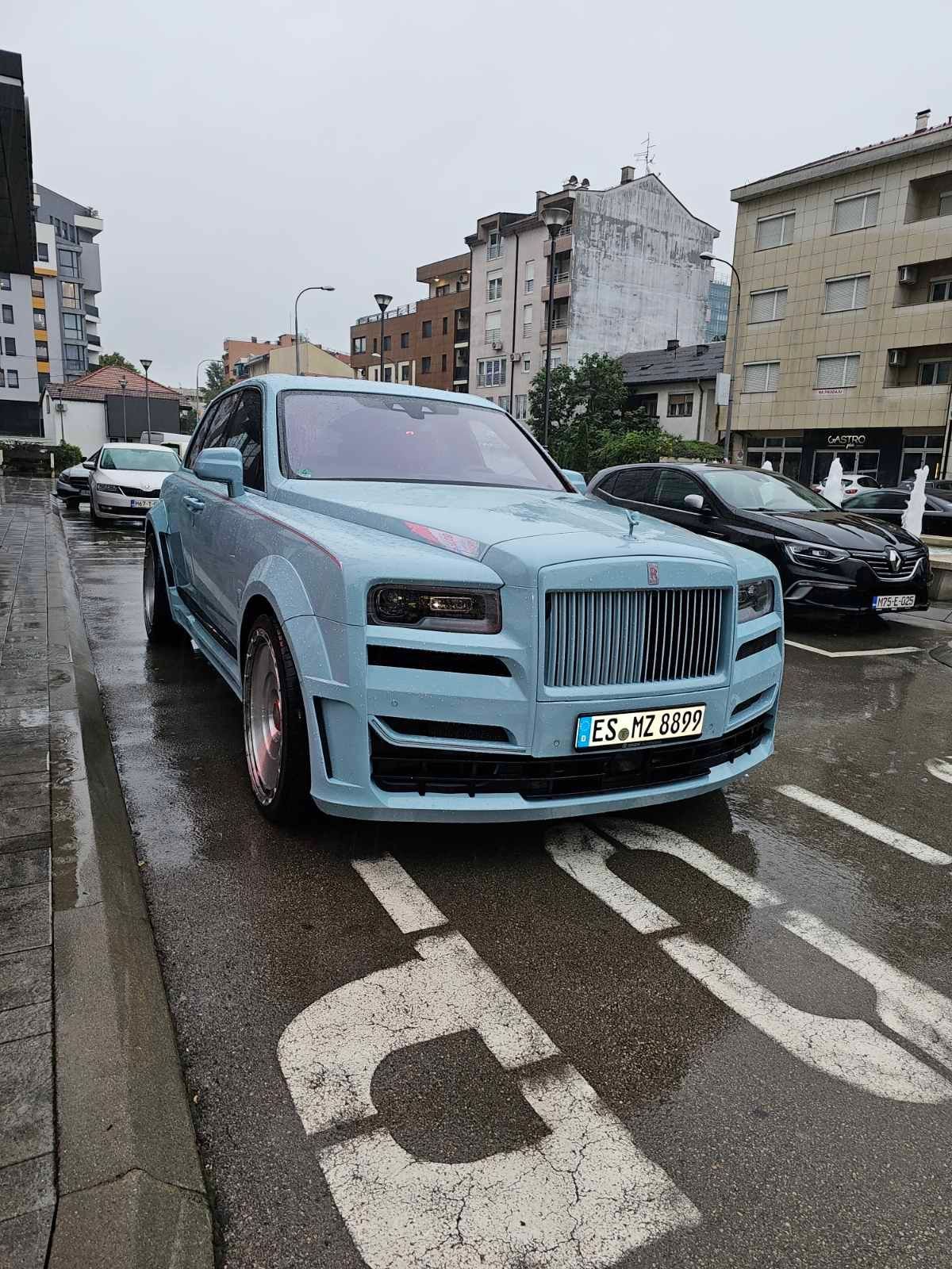 Light blue luxury car parked on wet street with surrounding buildings.