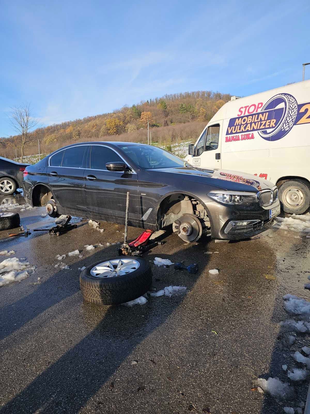 Car with removed wheel being serviced by a mobile vulcanizer van in an outdoor setting.