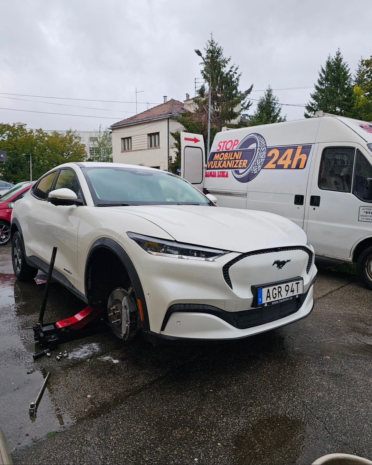 White car with a wheel removed, parked near a service van on a rainy day.