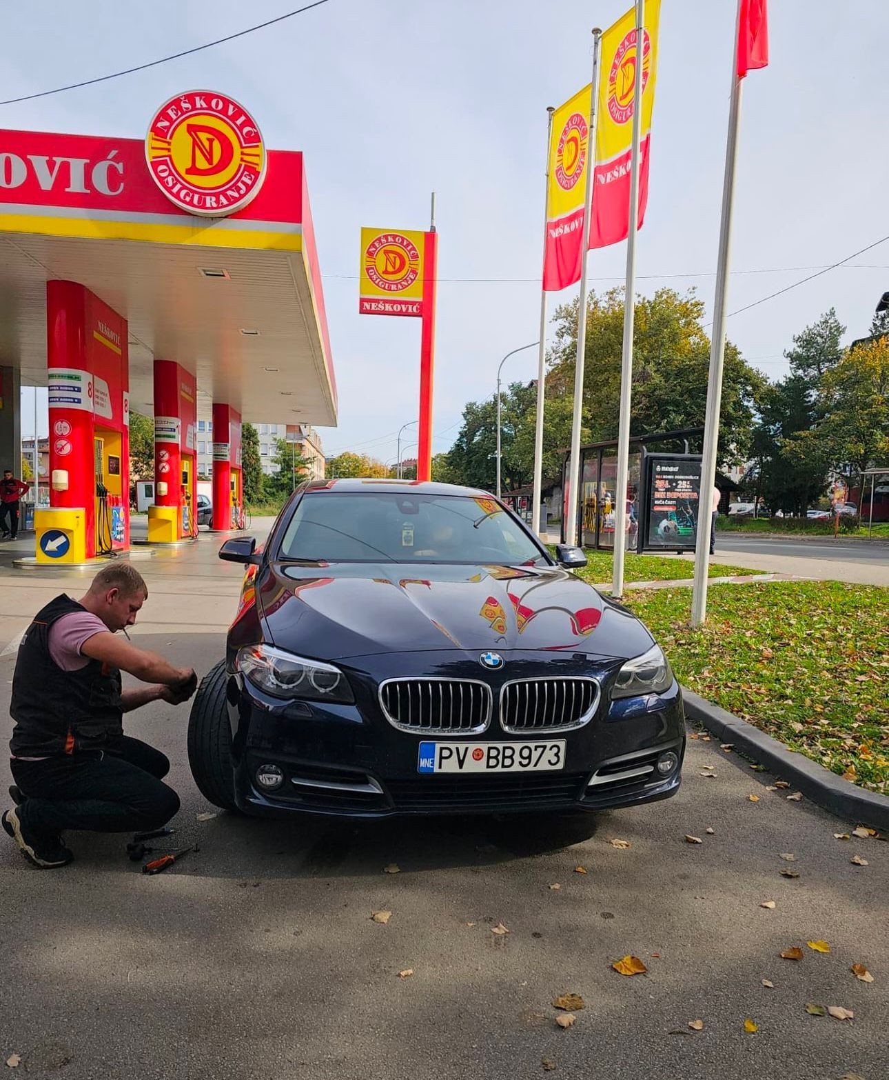 Black BMW car parked at a gas station with bright red and yellow signage and flags.